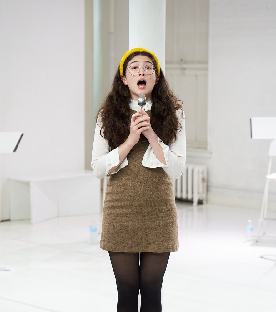 Photo of actor, Haley Wong, acting on stage. Performing a theater piece holding a spoon as a microphone. Her hair is messy. She is wearing a yellow puffy headband.